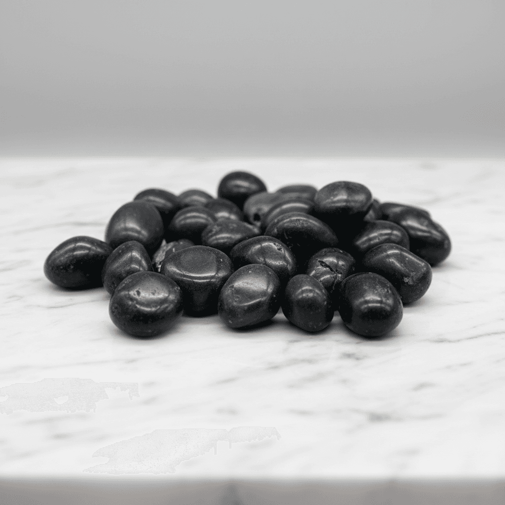 Present a professional studio environment with a minimalist aesthetic: Agni Manitite Tumbled Stones neatly arranged on a pristine white marble slab with subtle natural veining. Employ softbox lighting softened with diffusion to evenly illuminate the stones