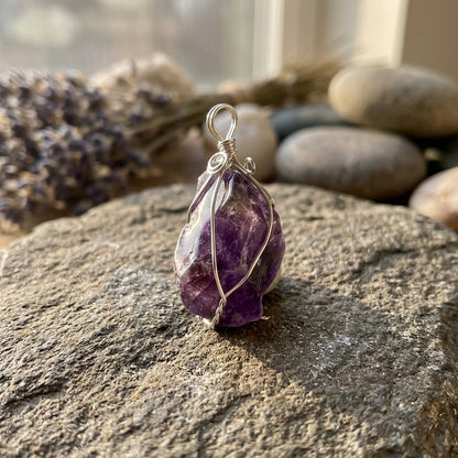 Close-up of a delicate amethyst wire wrapped pendant on a natural stone surface, illuminated by soft natural light highlighting its violet hue.