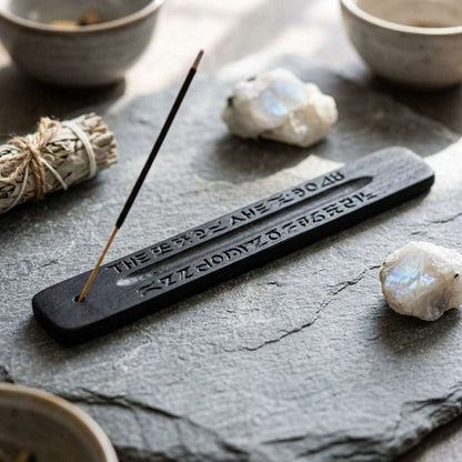 Detailed view of small black ash catcher with carved design and incense hole, placed on stone in a ritual setting with spiritual tools.