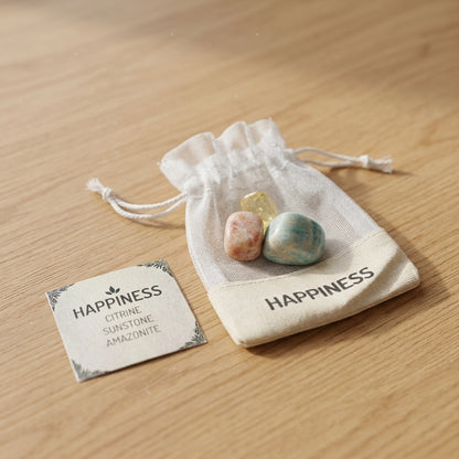 Close-up of a tiny Happiness Crystal Kit with Citrine, Sunstone, and Amazonite stones on a wooden surface, with a blurred 'HAPPINESS' bag and info card in the background.