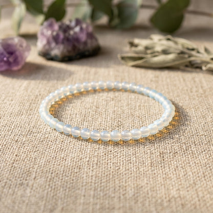 Opalite bracelet, 4mm, placed on linen fabric with a soft focus, illuminated by warm light, and surrounded by crystals and foliage for a spiritual ambiance.