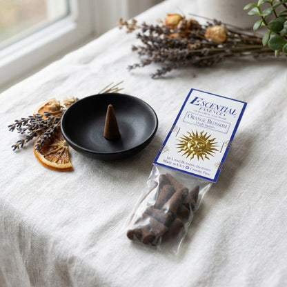 Serene scene of a small orange blossom incense cone on a black ceramic dish, illuminated softly against a minimal background.