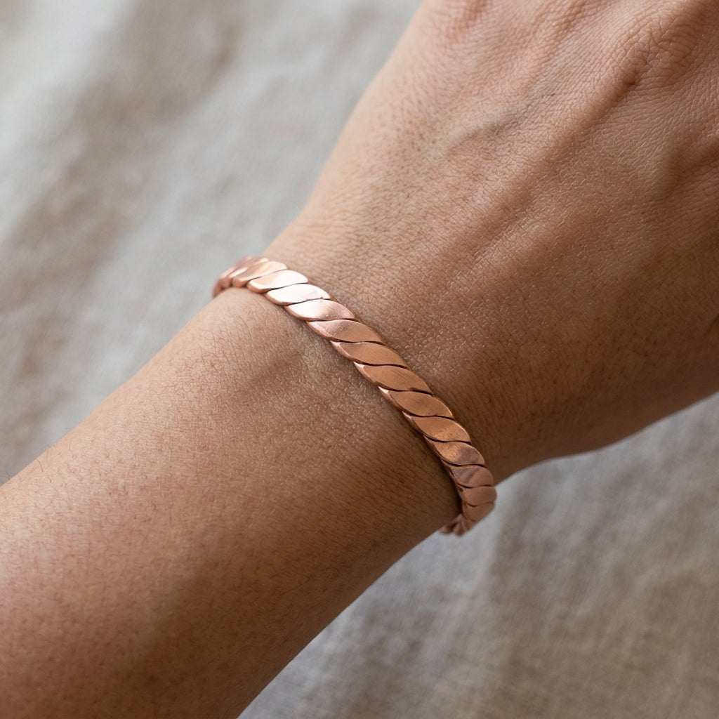 Photorealistic flat lay of a petite braided copper cuff bracelet on textured fabric, detailed craftsmanship visible with a small hand for scale.
