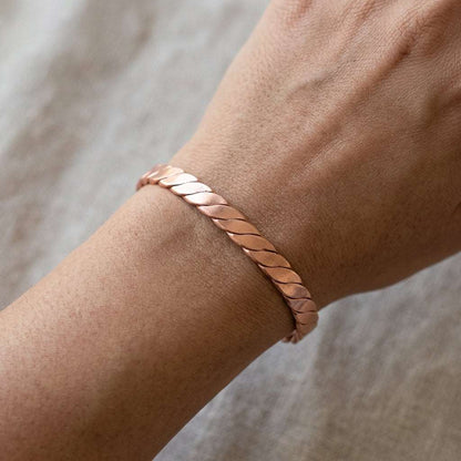 Photorealistic flat lay of a petite braided copper cuff bracelet on textured fabric, detailed craftsmanship visible with a small hand for scale.