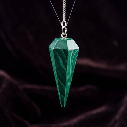 Create a high-resolution image of a 6-Sided Malachite Pendulum, 9 inches in length, suspended elegantly in a softly lit mystical setting. Use a diffused daylight studio light to highlight the rich, deep green banded patterns of the malachite's polished sur