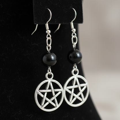 A close-up, hyper-realistic studio shot of Black Onyx Pentagram earrings displayed on a sleek, dark velvet background. The lighting is softbox diffused daylight, highlighting the glossy, polished black onyx beads and the metallic pewter charms with subtle 