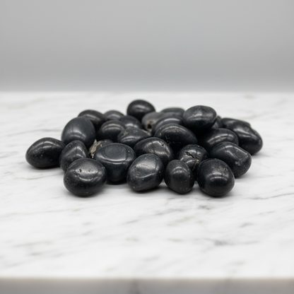 Present a professional studio environment with a minimalist aesthetic: Agni Manitite Tumbled Stones neatly arranged on a pristine white marble slab with subtle natural veining. Employ softbox lighting softened with diffusion to evenly illuminate the stones