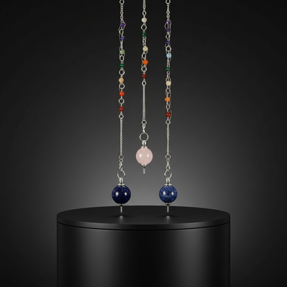 A sleek, modern sacred space arranged within a studio environment, illuminated with rim lighting that highlights the glossy texture of the chakra balls. Positioned on a matte black pedestal, the 7 Chakra Assorted Ball Pendulum is centered in the frame, per