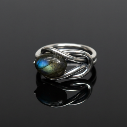 Create a highly detailed, studio-quality image of a size 12 sterling silver Labradorite ring, positioned on a sleek, reflective black surface. Use a softbox lighting setup to produce gentle, diffused daylight illumination that accentuates the iridescent fl