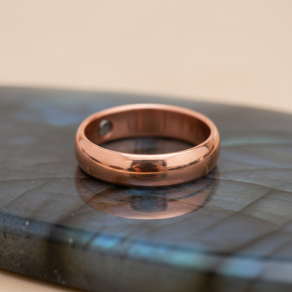 A close-up, high-resolution image of a copper magnetic solid band ring size 9, expertly displayed on a minimalist, reflective gemstone surface. The ring is illuminated with softbox lighting that highlights its rich, warm copper hue and the subtle texture o