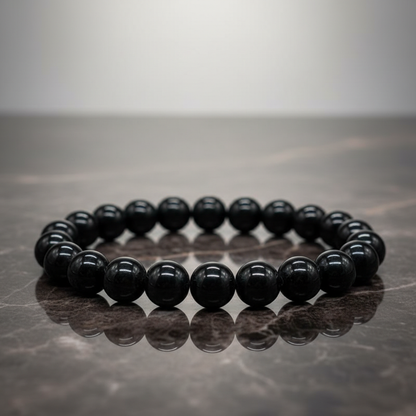 A close-up shot of the Jet Gemstone Bracelet, 8mm beads, elegantly displayed on a reflective dark marble surface under softbox lighting that enhances the smooth, glossy texture of the jet stones. The camera angle is straight-on with a focus on the uniformi