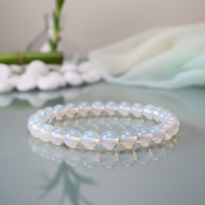 Capture a close-up, macro shot of the Opalite Beaded Bracelet, 8mm, elegantly draped around a reflective glass surface in a professional studio setting. Use softbox lighting to create gentle, diffused illumination that highlights the bracelet’s ethereal tr