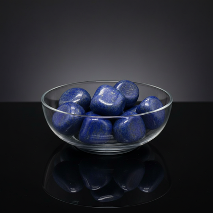 Create a high-resolution image of a luxurious, professional studio setup focused on a transparent, shallow glass bowl filled with roughly 6 to 11 natural Lapis Lazuli free shape stones, weighing approximately 1kg, arranged organically on a sleek, reflectiv