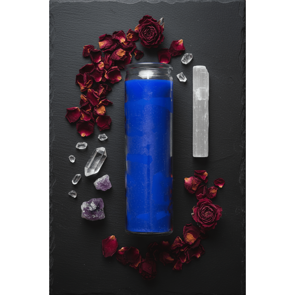 Flat lay of blue 7-day jar candle on dark slate with dried rose petals, selenite wand, and crystals