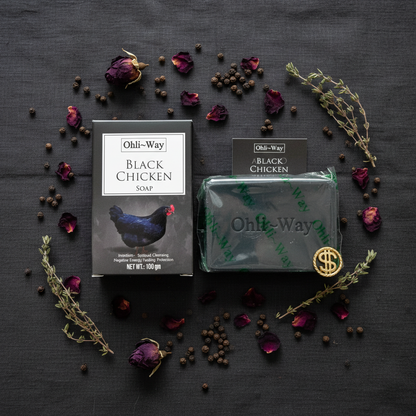 Ohli-Way Black Chicken Soap box, wrapped bar, insert card, and gold coin charm in overhead flat lay on dark linen with rose petals and herbs