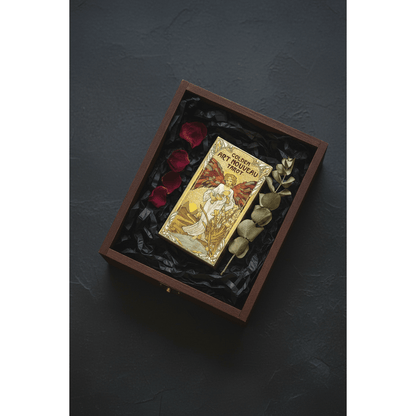 Golden Art Nouveau Tarot box nestled in black tissue paper inside an open dark gift box with dried rose petals and eucalyptus, gold foil catching soft warm light