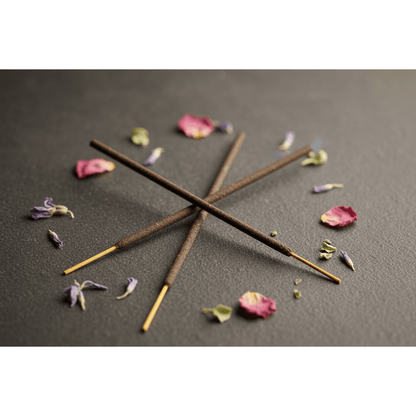 Two Sonavi 7 Chakra incense sticks crossed on dark stone with scattered dried flower petals and herb fragments.