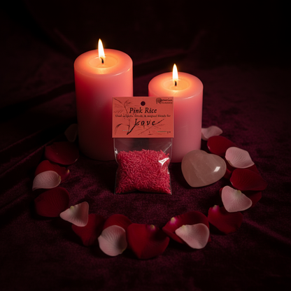 1oz Pink Infused Love Rice bag centered in a rose petal circle on dark velvet, flanked by pink pillar candles and heart-shaped rose quartz