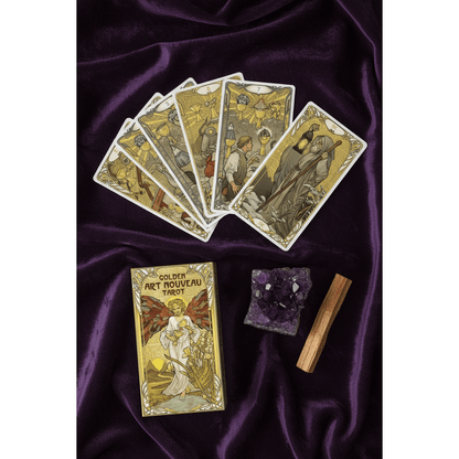 Golden Art Nouveau Tarot cards fanned face-up on dark velvet showing gold foil Art Nouveau illustrations, with amethyst cluster and palo santo beside the box