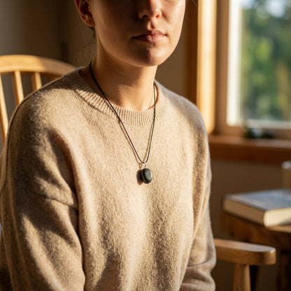 Tiny shungite pendant hanging from a thin cord in warm natural light, emphasizing dark grey tones and textured surface.