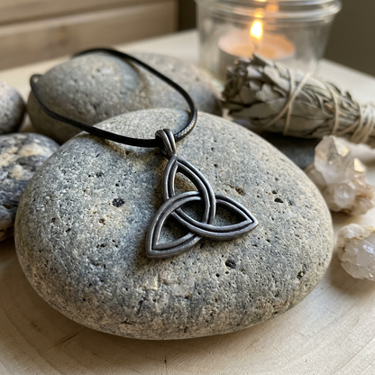 Photorealistic close-up of a tiny pewter Triskele amulet resting on a smooth stone in a sacred, softly lit space with herbs and sage.